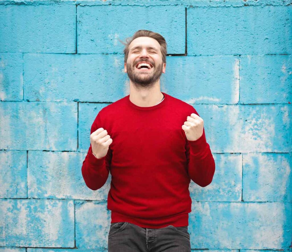 This holistic process will help you reach your authentic self and then grow from there. Image of a man in a red sweatshirt in front of a blue wall with his eyes closed and hands in fists smiling in victory.